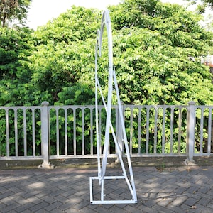 May include: White metal archway with a rounded top, standing on a gray brick surface. The archway is made of metal bars and has a simple, modern design.