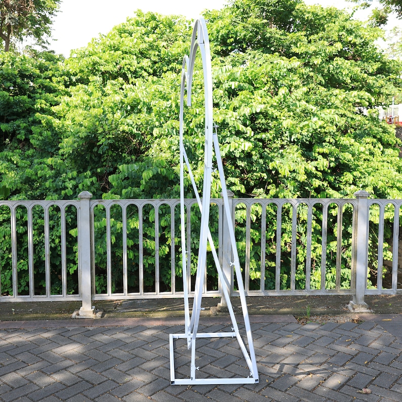 May include: White metal archway with a rounded top, standing on a gray brick surface. The archway is made of metal bars and has a simple, modern design.