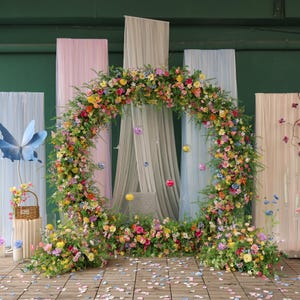 Pode incluir: Um arco floral circular adornado com uma vibrante variedade de flores coloridas e vegetação. O arco é colocado em frente a um pano de fundo de painéis de tecido em tons pastel e elementos decorativos, criando um cenário caprichoso e romântico. Pétalas espalhadas no chão de madeira.