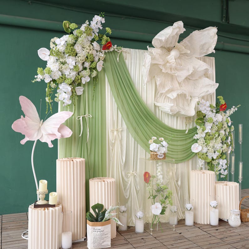 May include: A decorative backdrop featuring a large paper flower, draped fabric, and floral arrangements. A large pink butterfly decoration is on the left. Beige cylindrical pedestals and candles are in the foreground. The backdrop is green and white.