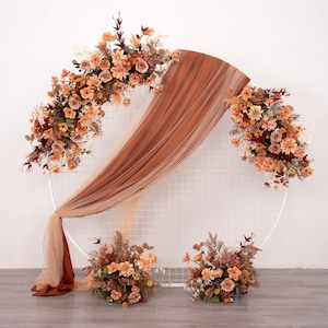 Puede incluir: Un arco circular de metal blanco decorado con tela de seda marrón y naranja y flores artificiales. Las flores son en tonos marrones, naranjas y rojos.