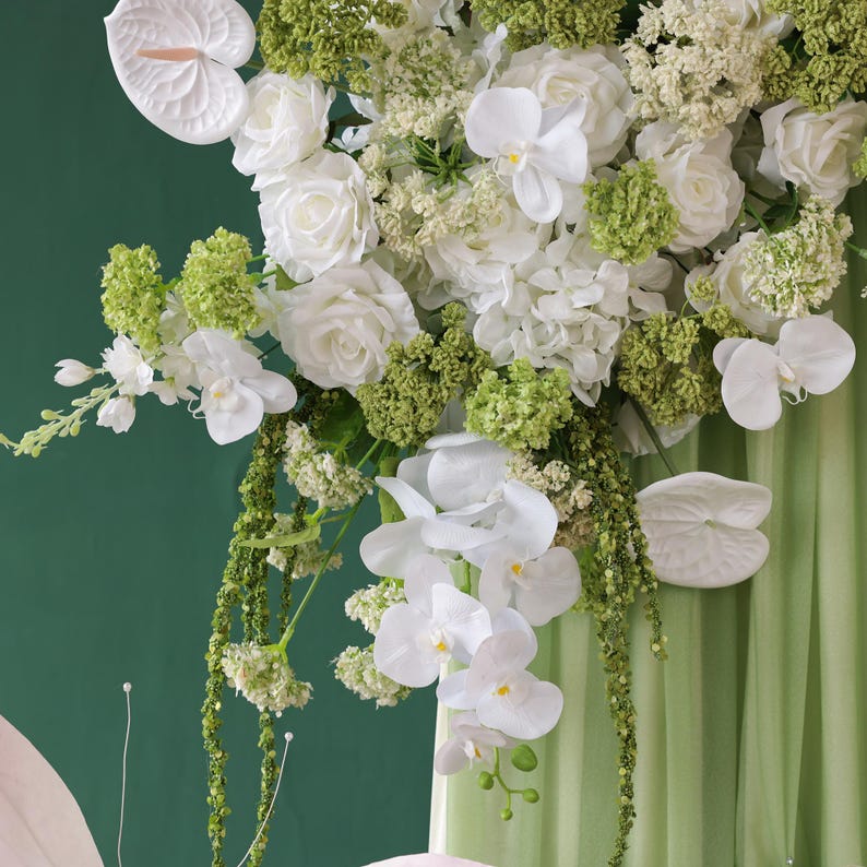 May include: A lush floral arrangement featuring white roses, orchids, and green accents. The bouquet includes cascading greenery and is set against a soft green backdrop, creating a fresh and elegant display.