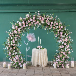 May include: A floral arch made of pink and white flowers, with a white lace tablecloth and candles on a table in front of it.