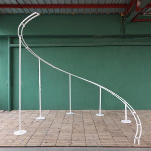 以下が含まれることがあります： 白い金属製のアーチ型の背景スタンドで、湾曲した支柱が付いています。スタンドは、湾曲した形状を作るために、複数のセクションで構成されています。スタンドは緑色の壁に設置されています。