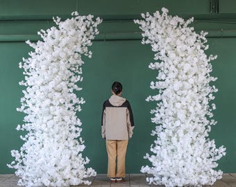 May include: Two large white flower arrangements create a backdrop for a person standing in the middle. The flowers are arranged in a tall, vertical shape, resembling a pair of columns.