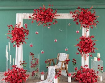 May include: A wedding arch display featuring white fabric drapes adorned with large arrangements of vibrant red roses. The setup includes a wooden chair with a white runner, hanging glass orbs, and floor-standing floral clusters.