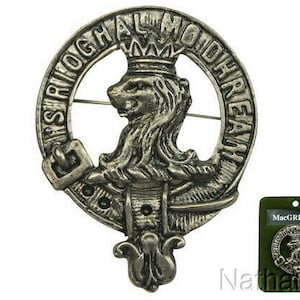 May include: A silver-toned clan crest badge featuring a lion rampant within a belt inscribed with Gaelic text. The badge has a pin on the back. A green card with the name "MacGREGOR" is also visible.
