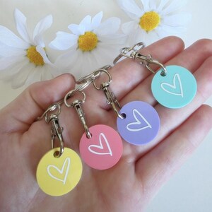 Könnte beinhalten: In einer Hand werden vier runde Schlüsselanhänger in Gelb, Rosa, Lila und Türkis gehalten. Jeder Anhänger hat ein weißes Herz-Design. Die Schlüsselanhänger sind an silbernen Metallverschlüssen und Schlüsselringen befestigt. Weiße Gänseblümchen sind im Hintergrund.
