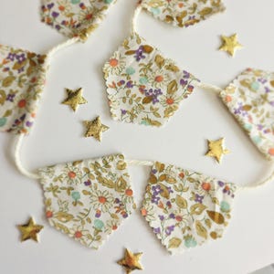 May include: A decorative banner made of floral fabric, strung on a cream-coloured cord, with gold star-shaped decorations. The fabric has a white base with a pattern of small flowers in purple, orange, and green. The banner is suitable for celebrations.
