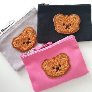 Könnte beinhalten: Drei kleine Reißverschlusstaschen mit braunen, flauschigen Bären-Applikationen. Die Taschen sind grau, schwarz und rosa.