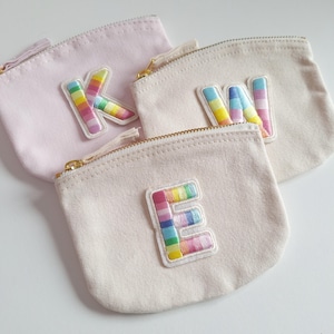 May include: Three small zippered pouches made of beige canvas with rainbow embroidered letters K, W, and E. The pouches are lined up in a row, with the K pouch on the left, the W pouch in the middle, and the E pouch on the right.
