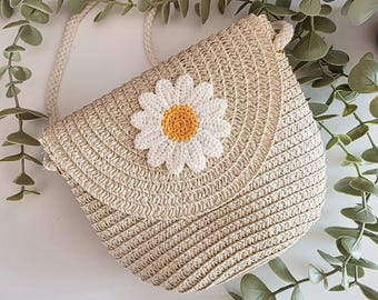 May include: A beige woven straw crossbody bag featuring a circular flap closure. The center of the flap is decorated with a large, crocheted daisy flower applique with white petals and a textured yellow center. It includes a braided rope strap.