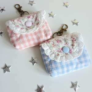 May include: Two small, quilted fabric pouches with keychains. One is pink gingham with a floral flap and white lace trim. The other is blue gingham with a floral flap and white lace trim. Both have a button closure.
