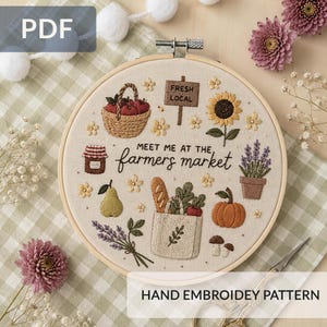 May include: An embroidery hoop featuring a cream fabric with stitched designs of a basket of apples, a jar, a pear, a tote bag with bread, a pumpkin, mushrooms, lavender, a sunflower, and a sign reading "MEET ME AT THE farmers market".