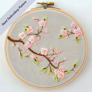 May include: A hand-embroidered floral design on grey fabric, set in a wooden hoop. The embroidery depicts pink cherry blossoms with yellow centres, green leaves, and brown branches. The text "Hand Embroidery Pattern" is visible.