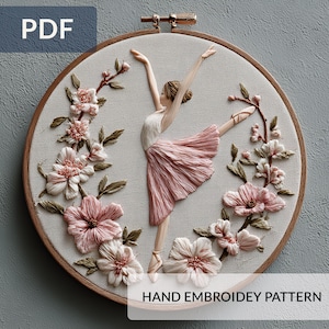 Könnte beinhalten: Ein handgesticktes Stickrahmen-Kunstwerk mit einer Ballerina in einem rosa Tutu und floralen Akzenten. Das Design zeigt eine Tänzerin mit erhobenen Armen, umgeben von rosa und cremefarbenen Blumen und grünen Blättern. Der Text "HAND EMBROIDERY PATTERN" ist sichtbar.