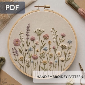 Può includere: Un telaio da ricamo circolare in legno contenente un pannello in tessuto beige con un disegno floreale cucito a mano che presenta vari fiori di campo nelle tonalità del rosa, blu, giallo e bianco, con il testo HAND EMBROIDEY PATTERN in basso.