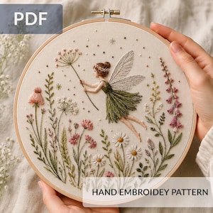 May include: A circular wooden embroidery hoop containing a fabric panel with a hand-stitched design of a fairy with wings wearing a green dress, surrounded by various embroidered wildflowers, stems, and leaves in shades of pink, white, and green.