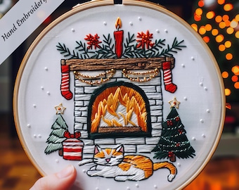 May include: Hand-embroidered hoop art featuring a cozy fireplace scene with a lit candle, garland, stockings, and a cat resting in front. The fireplace is decorated with brickwork and a roaring fire. Two small Christmas trees flank the fireplace.