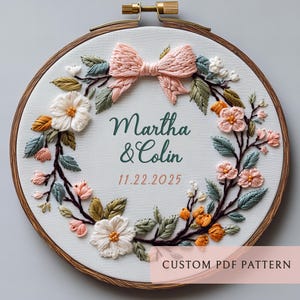 Könnte beinhalten: Gestickter Stickrahmen mit floralem Kranz und Pfirsichschleife. Die Namen "Martha & Colin" und das Datum "11.22.2025" sind grün gestickt. Der Holzrahmen hält das Design, mit dem Text "CUSTOM PDF PATTERN" unten.