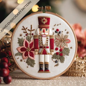 Puede incluir: Adorno navideño bordado a mano con un diseño de soldado cascanueces. El bordado está colocado en un aro de madera e incluye colores rojo, blanco y dorado. El texto "HAND EMBROIDERY PATTERN" está impreso en la esquina superior izquierda.