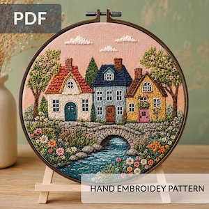 Pode incluir: Um bastidor de bordado circular contendo uma cena costurada à mão de três casas com telhados vermelhos, azuis e amarelos atrás de uma ponte de pedra sobre um riacho azul. O design apresenta árvores verdes, flores coloridas e nuvens brancas.