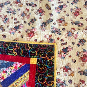May include: A handmade quilt featuring a cream-colored background with repeating patterns of teddy bears, American flags, airplanes, and cars. The quilt has a colorful border with a black background and rainbow-colored circles, and a patchwork design.