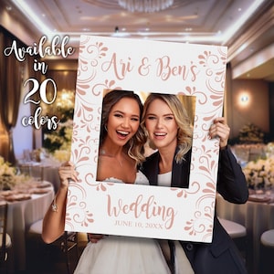 May include: A white photo booth prop with a rose gold frame and floral design. The frame has a cut-out for a face and the text "Ari & Ben's Wedding June 10, 20XX".