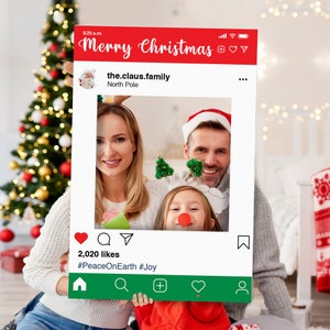 May include: A red and white photo booth prop with a Christmas theme. The prop features a mock Instagram post with the text "Merry Christmas" and "the.claus.family North Pole". The post also includes a photo of a family with a child wearing reindeer antlers.