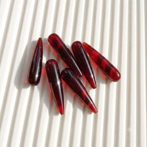 May include: A collection of six elongated, teardrop-shaped beads in a deep, translucent red hue. Each bead features a smooth, polished surface with a tapered point at one end and a rounded top, reflecting light with a glossy, glass-like finish.