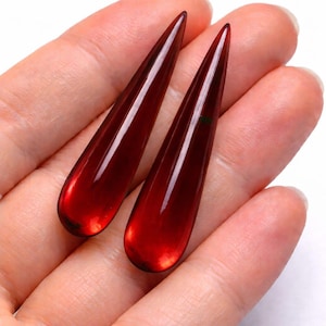 Puede incluir: Dos colgantes alargados en forma de lágrima, de color rojo intenso. Los colgantes son lisos, brillantes y parecen estar hechos de un material translúcido. Se sostienen en una mano, mostrando su color vibrante y su diseño elegante.
