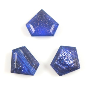 Op de afbeelding: Drie blauwe edelsteen cabochons met een vijfhoekige vorm. De edelstenen hebben een gespikkelde, glinsterende uitstraling, met donkere en lichte blauwtinten. De edelstenen liggen op een witte achtergrond.
