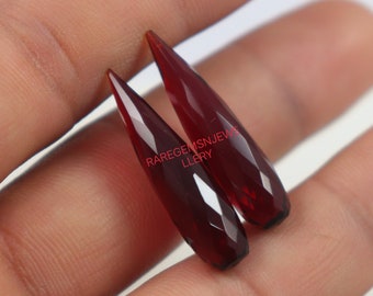 Può includere: Due punte di pietre preziose sfaccettate rosso scuro, anche note come forme a goccia o a proiettile, sono mostrate su una mano. Le pietre preziose misurano circa 5 cm di lunghezza. Il testo "RAREGEMS JEWELLERY" è visibile nell'immagine.