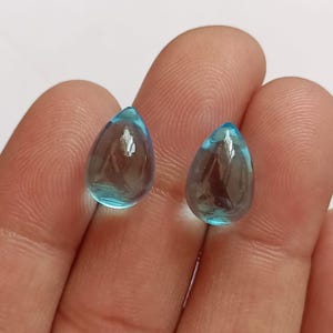 Pode incluir: Duas contas de vidro azul em forma de lágrima. As contas são transparentes e têm um leve brilho.