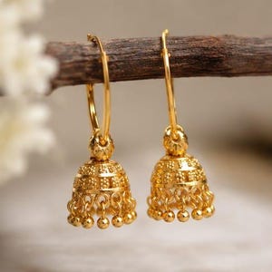 May include: A pair of gold hoop earrings featuring dangling bell-shaped jhumka charms. Each charm has a textured, granulated surface with small, round beads hanging from the bottom edge. The earrings are suspended from a thin, dark brown twig.