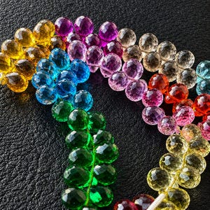 Peut inclure: Gros plan sur une collection de perles de pierres précieuses à facettes dans un arc-en-ciel de couleurs. Les perles sont disposées en rangées incurvées, présentant des nuances de jaune, de vert, de bleu, de violet et de rouge. Les perles sont enfilées, et le fond est noir.