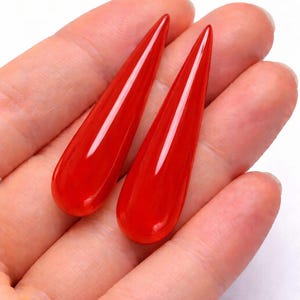 May include: Two vibrant red, teardrop-shaped pendants. The polished, elongated drops have a glossy finish, reflecting light. The jewelry pieces are displayed against a neutral background, highlighting their color and form. These are likely earrings or necklace pendants.