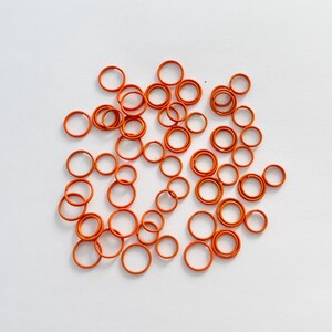 May include: A collection of small, orange, circular rubber rings scattered on a white surface. The rings vary in size, with some overlapping. The bright orange color provides a contrast against the white background.