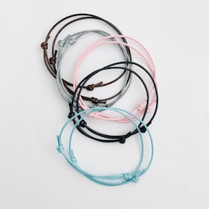 May include: Five colorful friendship bracelets in various colors, including light blue, pink, gray, brown, and black. The bracelets are made of thin cord and have a simple knot design.