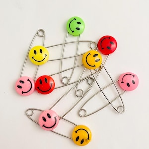 May include: A group of safety pins with smiley face charms in various colors, including yellow, red, green, pink, and orange.
