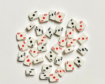 May include: A collection of white ceramic playing card charms with red and black suits. The charms are shaped like playing cards and have a hole in the top for stringing.