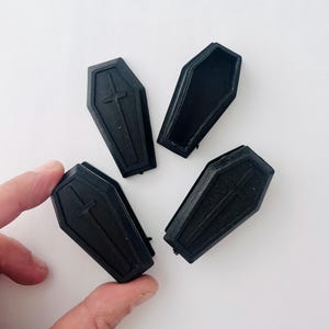 May include: Four black plastic coffin-shaped boxes with a cross design. The boxes are small and appear to be decorative.