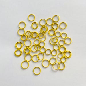 May include: A large group of yellow metal jump rings. The rings are scattered on a white surface.