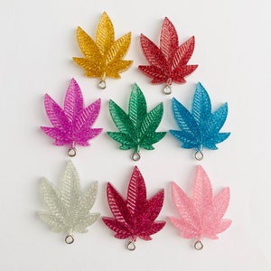 May include: A collection of glittery, cannabis leaf-shaped charms in various colors, including gold, red, purple, green, blue, silver, dark red, and pink. Each charm has a small metal loop for attaching to jewelry or other crafts.