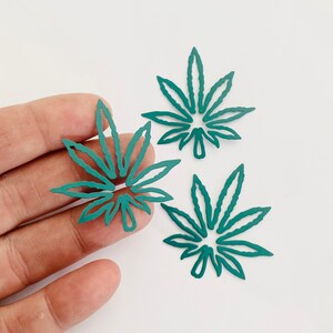 May include: Three teal green leaf-shaped cutouts, resembling marijuana leaves, are arranged on a white surface. The cutouts are made of a thin, metallic material.
