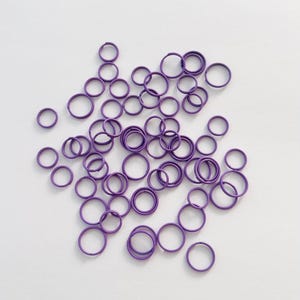 May include: A collection of small, purple metal rings scattered on a white surface. The rings are uniform in size and shape, with a smooth, matte finish. The image is a top-down view, with the rings taking up most of the frame.