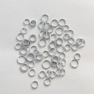 May include: A collection of small, white metal jump rings scattered on a white surface. The rings vary slightly in size and are open, ready for use in jewelry making or crafts. The lighting is bright, highlighting the smooth texture of the rings.