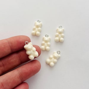 May include: Five small, off-white gummy bear charms with silver-colored loops for attaching to jewelry or crafts. The charms are arranged on a white surface, with one held by a hand.