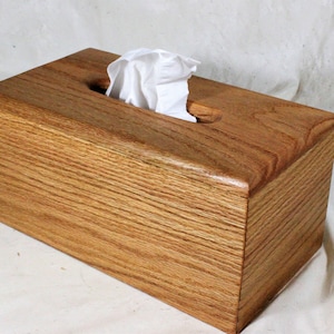 Może przedstawiać: Prostokątne drewniane pudełko na chusteczki z naturalnym wykończeniem słojów drewna. Pudełko ma gładką, wypolerowaną powierzchnię i prostokątny otwór na górze do dozowania chusteczek. Widoczne są białe chusteczki.