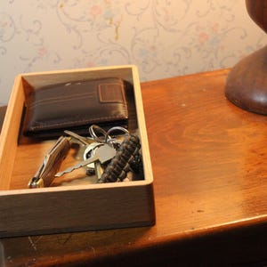 May include: A wooden tray holds a brown leather wallet, keys, and a pocket knife. The tray sits on a wooden surface next to a dark brown wooden lamp base. The background features floral wallpaper.